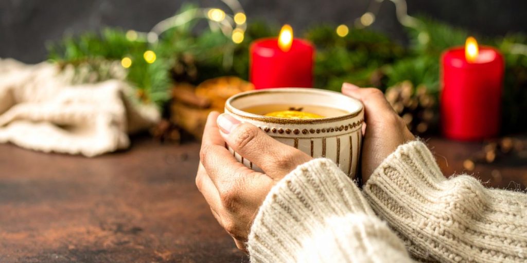 Hands holding a warm mug with holiday candles, pine branches, and soft lights in the background, symbolising comfort during emotional and physical stress.